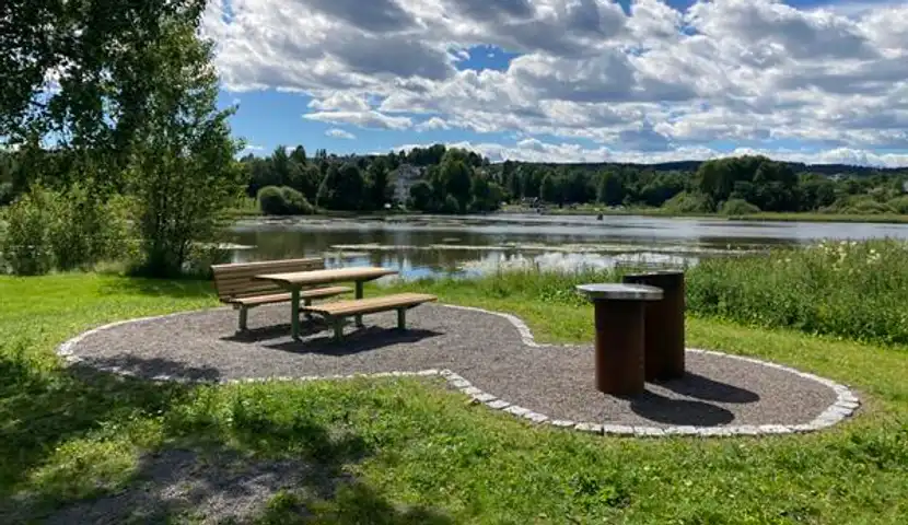 En park med en benk og en grill. Vann i bakgrunnen.